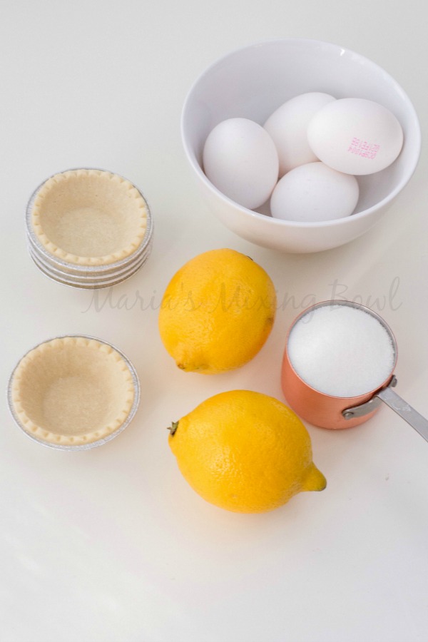 eggs in a bowl, sugar in a measuring cup, and mini pie shells and lemons sitting on a white counter