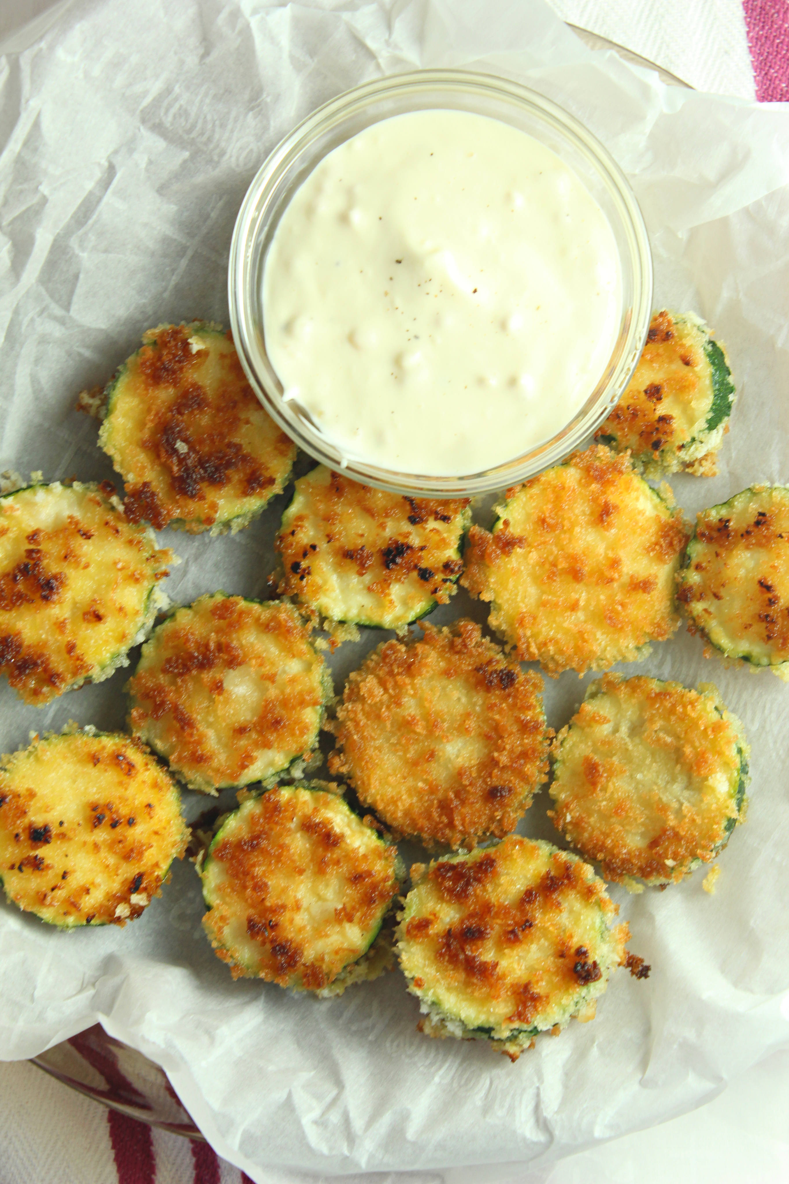 aioli sauce in a clear bowl with cooked zucchini rounds on white paper and white and pinkish red napkin 