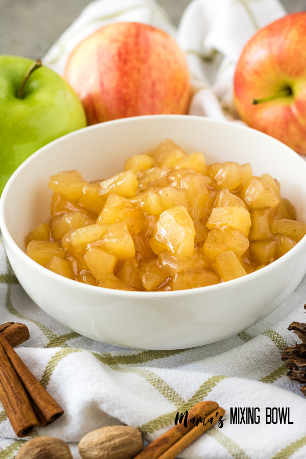 chopped canned apples in a white bowl with fresh apples on a white and brown striped towel