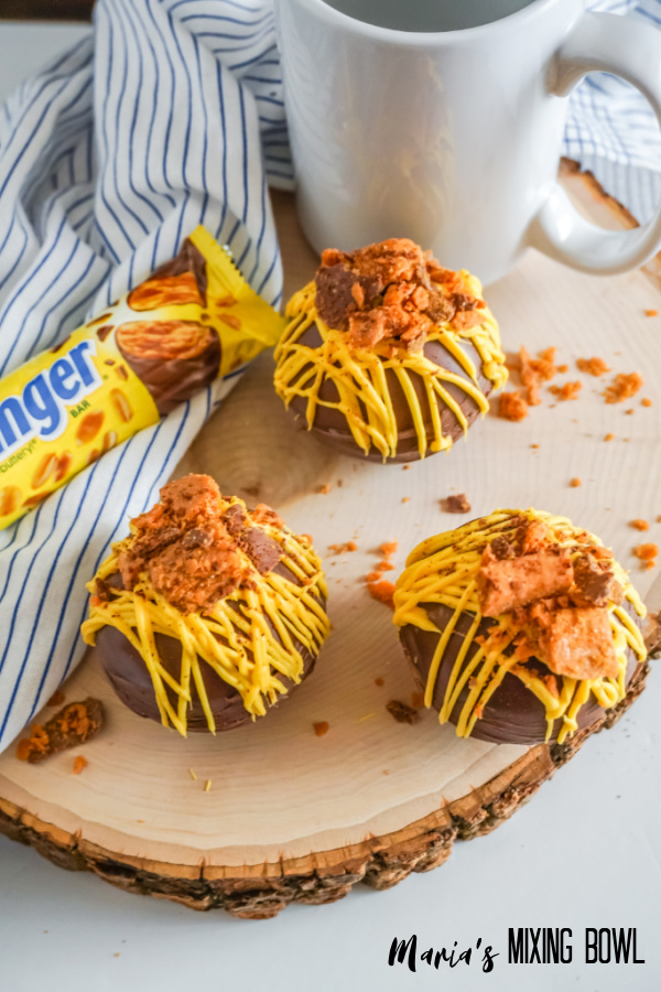 3 hot chocolate butterfinger bombs on a wood board with a white and blue napkin and white cup and candy bar