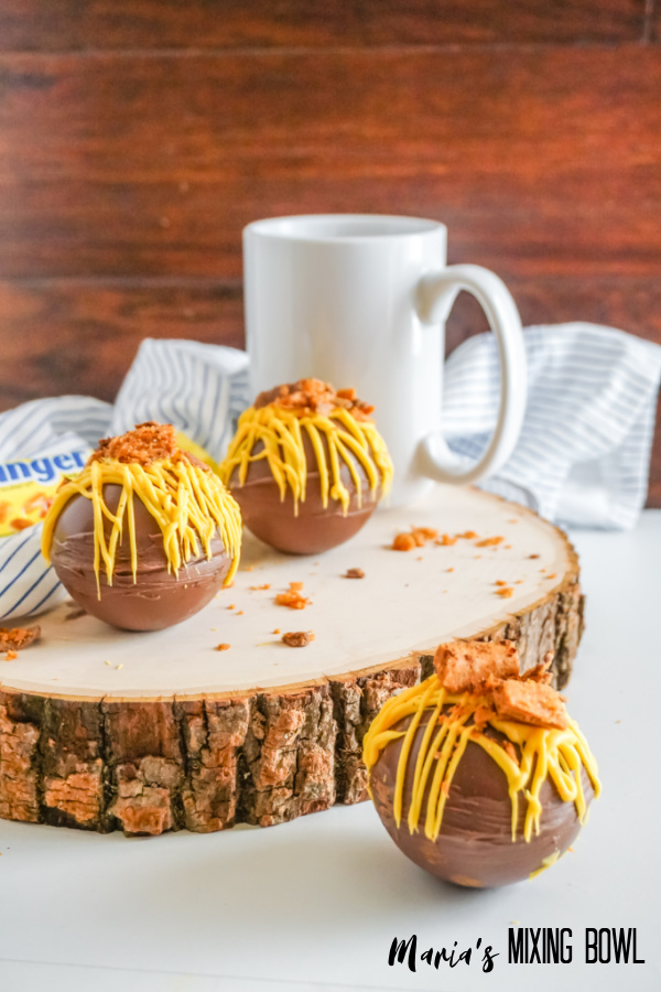 hot cocoa bombs wih yellow candy melt on a wooden board, white cup and white and blue strip napkin