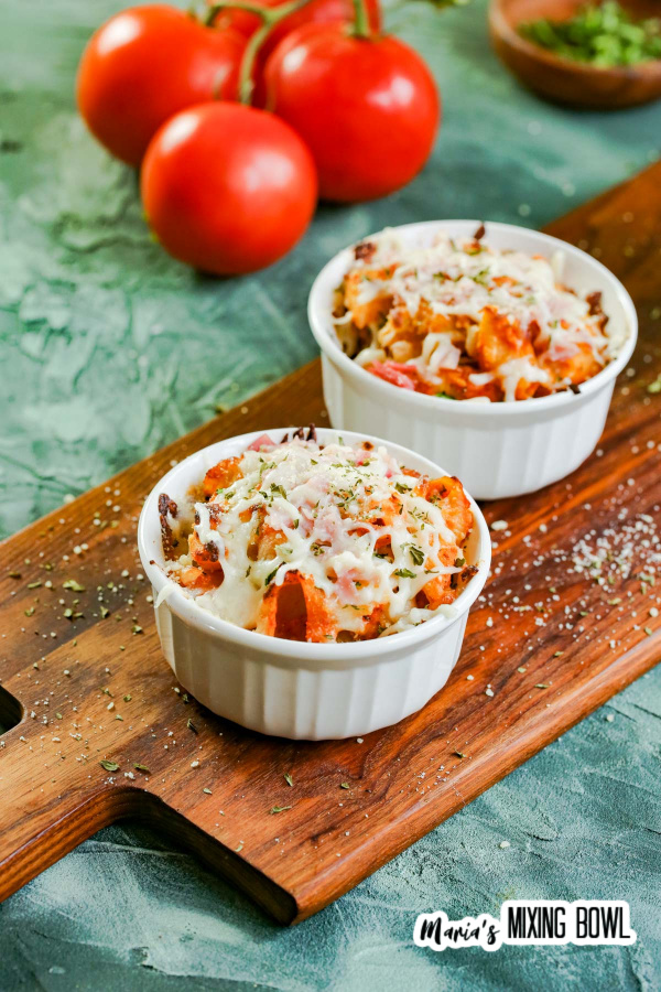 Two bowls of unstuffed shells casserole on wooden board with tomatoes in background