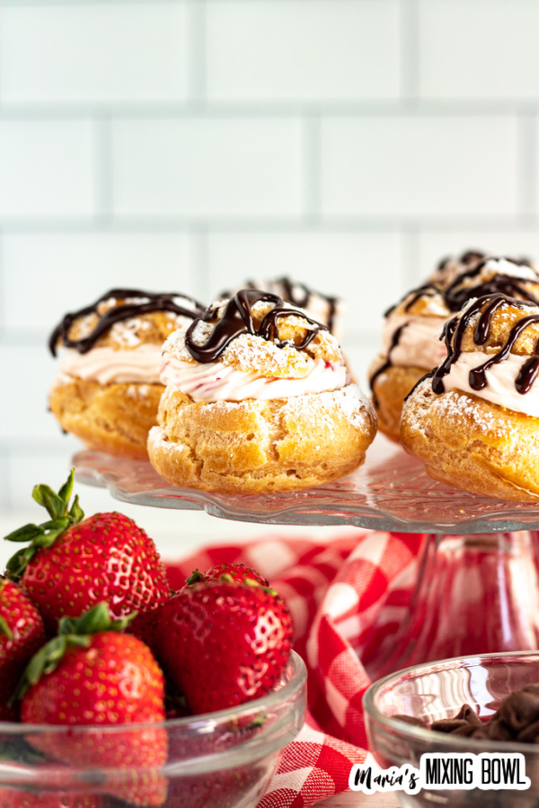 Cream puffs on glass cake dish with bowl of strawberries and bowl of chocolate chips in front