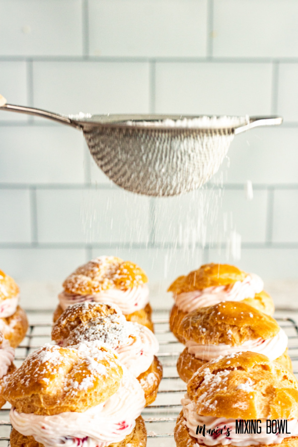 Sifter being used to sprinkle powdered sugar over strawberry cream puffs on wire rack