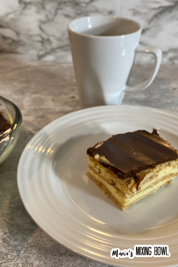 Slice of chocolate eclair cake on white plate with coffee mug in background