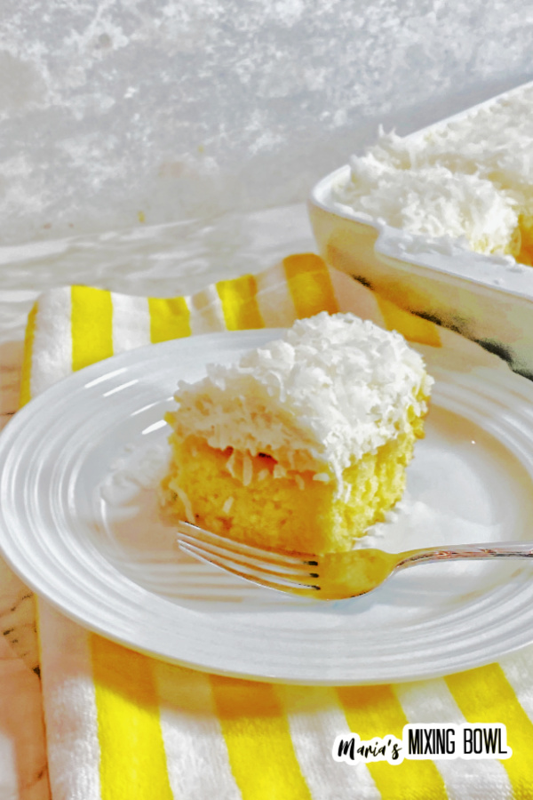 Slice of pineapple coconut poke cake with fork on white plate with more poke cake in baking dish in background