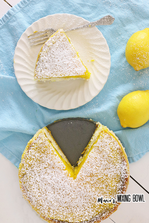 Overhead shot of slice of lemon bar cheesecake on white plate next to whole lemon bar cake