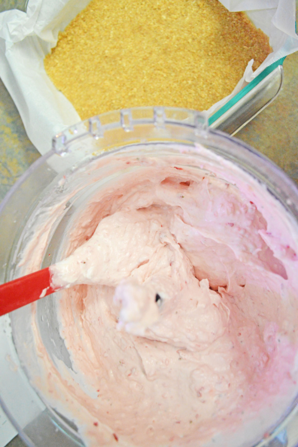 Overhead shot of spatulat stirring strawberry cheesecake mixture in food processor next to graham cracker crust