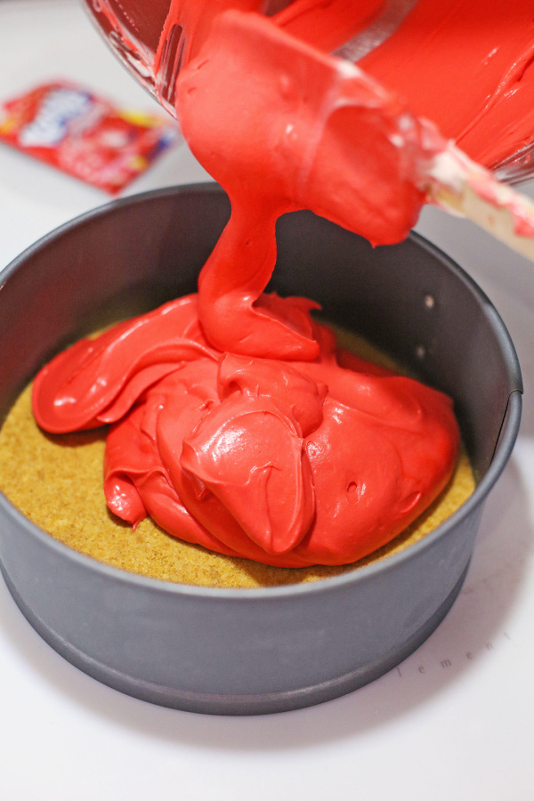 Red cheesecake batter being poured over graham cracker crust in springform pan