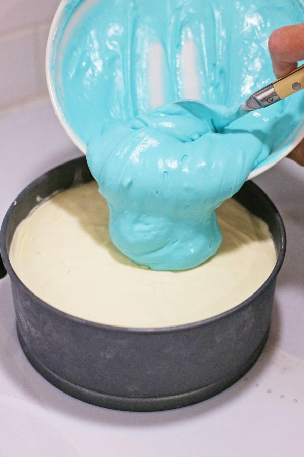 Blue cheesecake batter being poured over vanilla cheesecake batter in springform pan