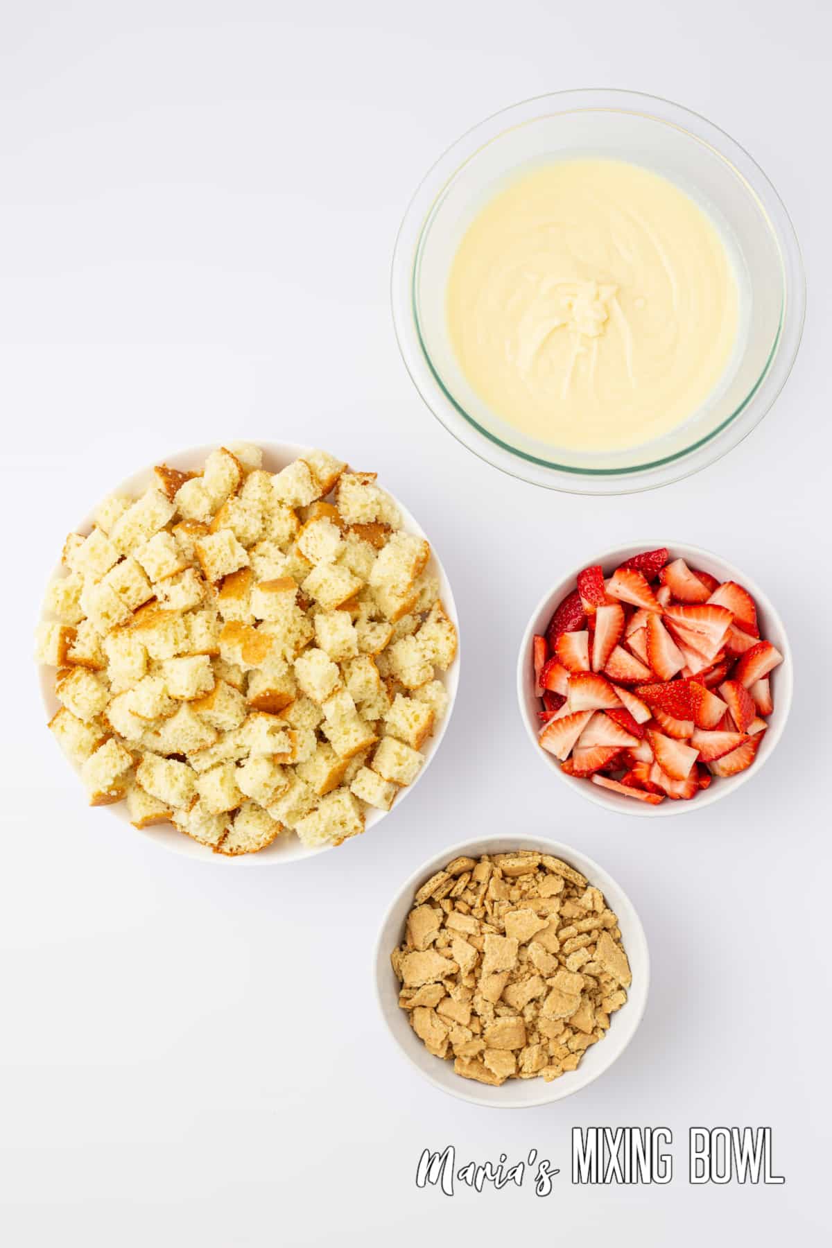 bowls of crushed graham crackers, sliced strawberries, pudding, and diced cake pieces on a white table