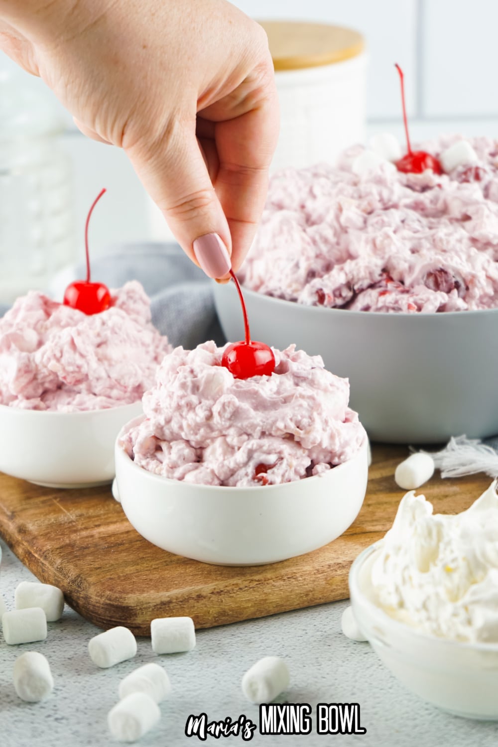 small white bowls filled with cherry fluff, with a women's fingers adding a cherry to the top