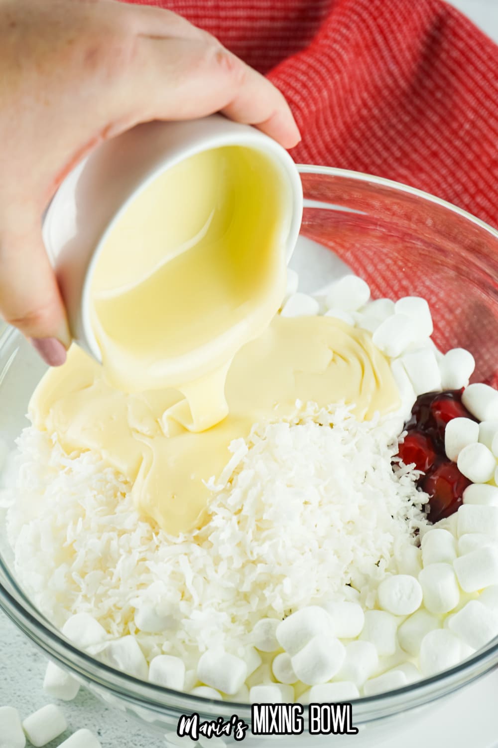 condensed milk being poured into a glass bowl with marshmallows, coconut, and cherries