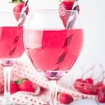 wine glass full of cupids cocktail with a strawberry on the rim with a black and white straw.