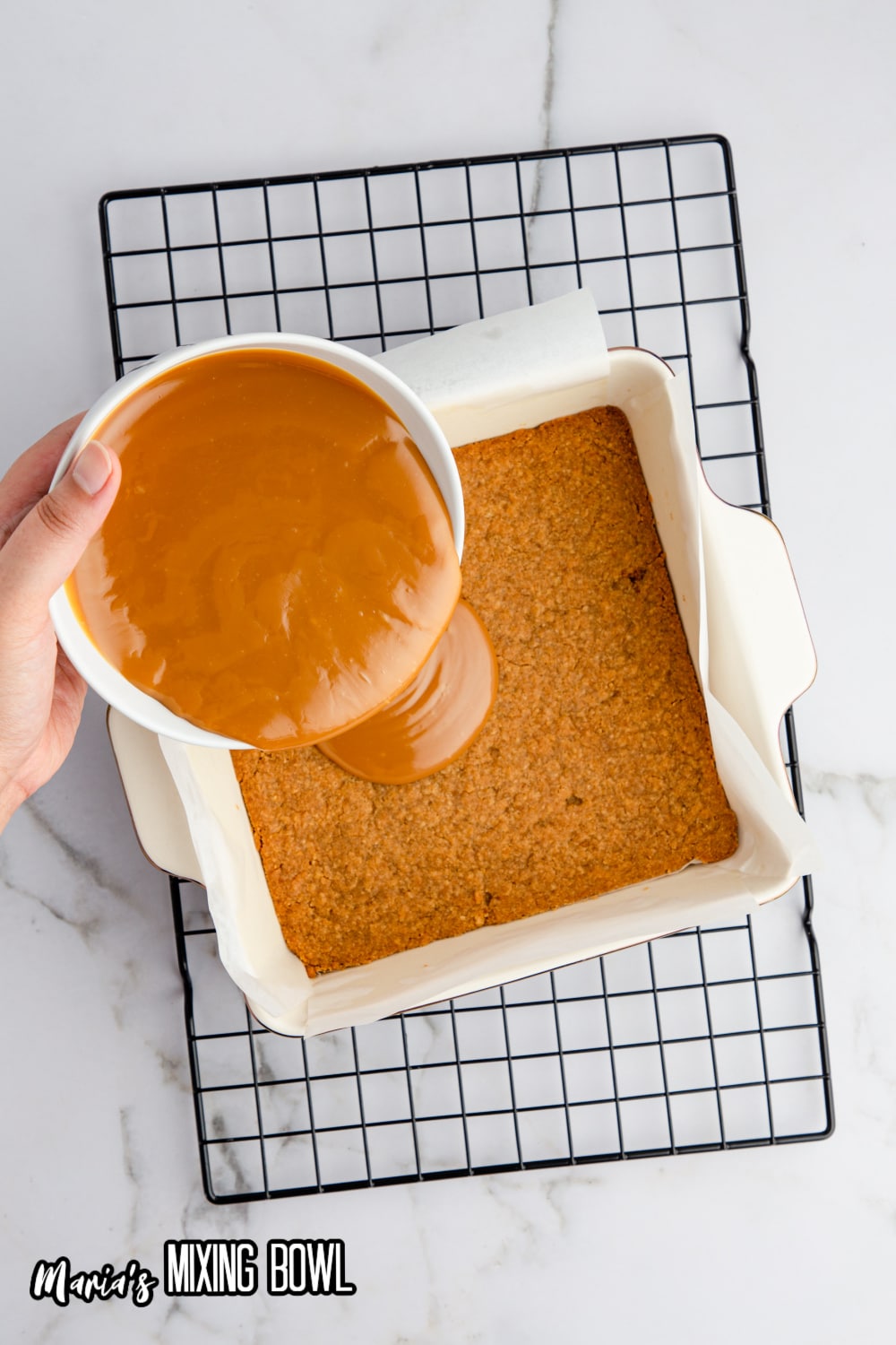 caramel being poured over a cookie crust in a parchment lined square baking dish