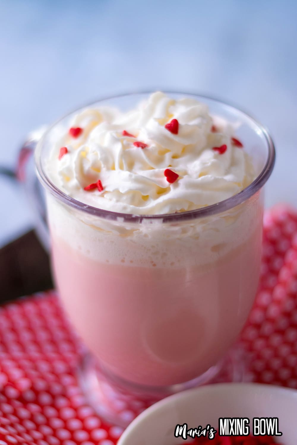 glass mug full of pink hot chocolate topped with whipped cream and red heart sprinkles