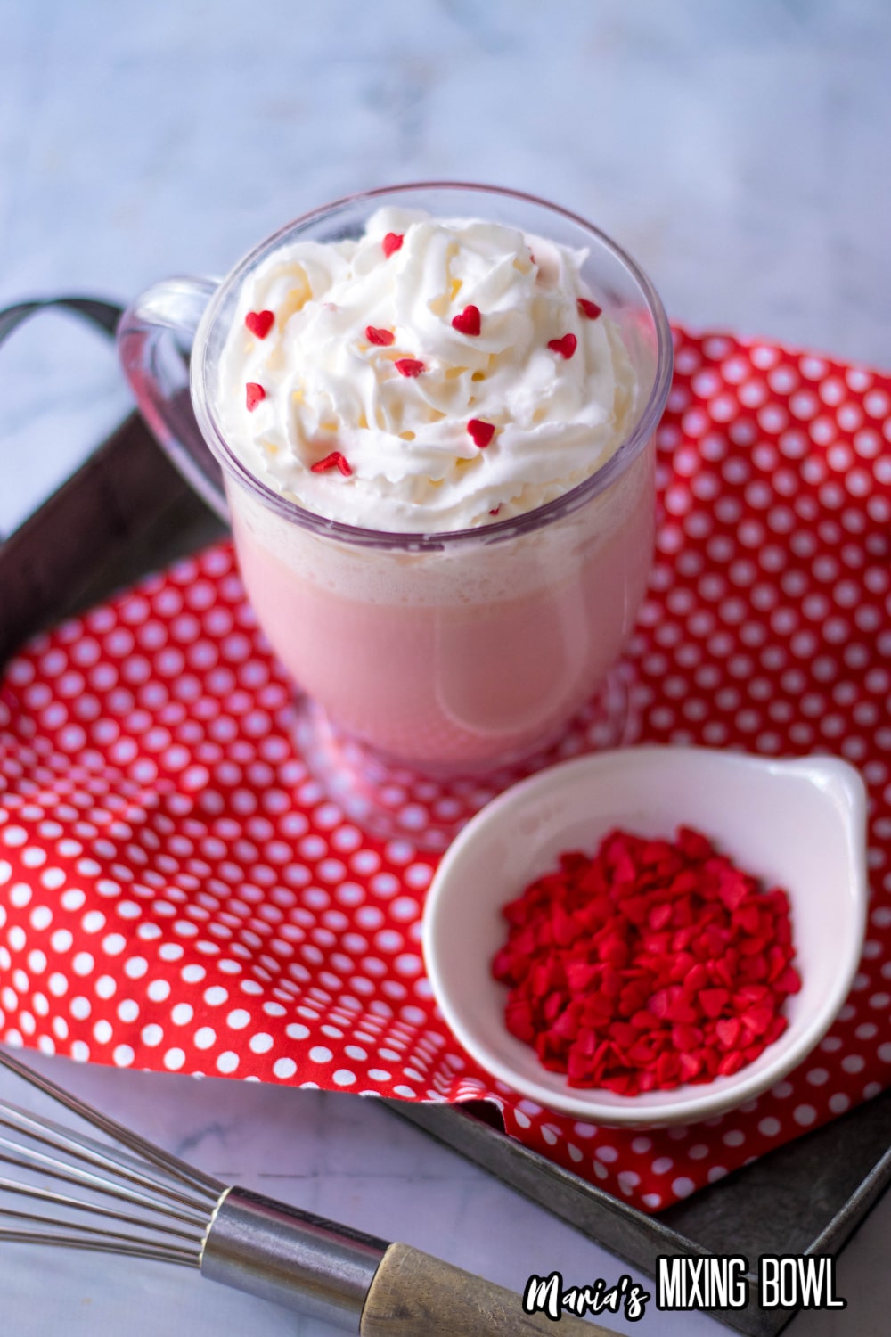 glass mug full of pink hot chocolate topped with whipped cream and red heart sprinkles