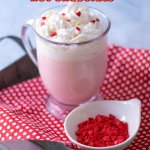glass mug full of pink hot chocolate topped with whipped cream and red heart sprinkles with the name in text for pinterest