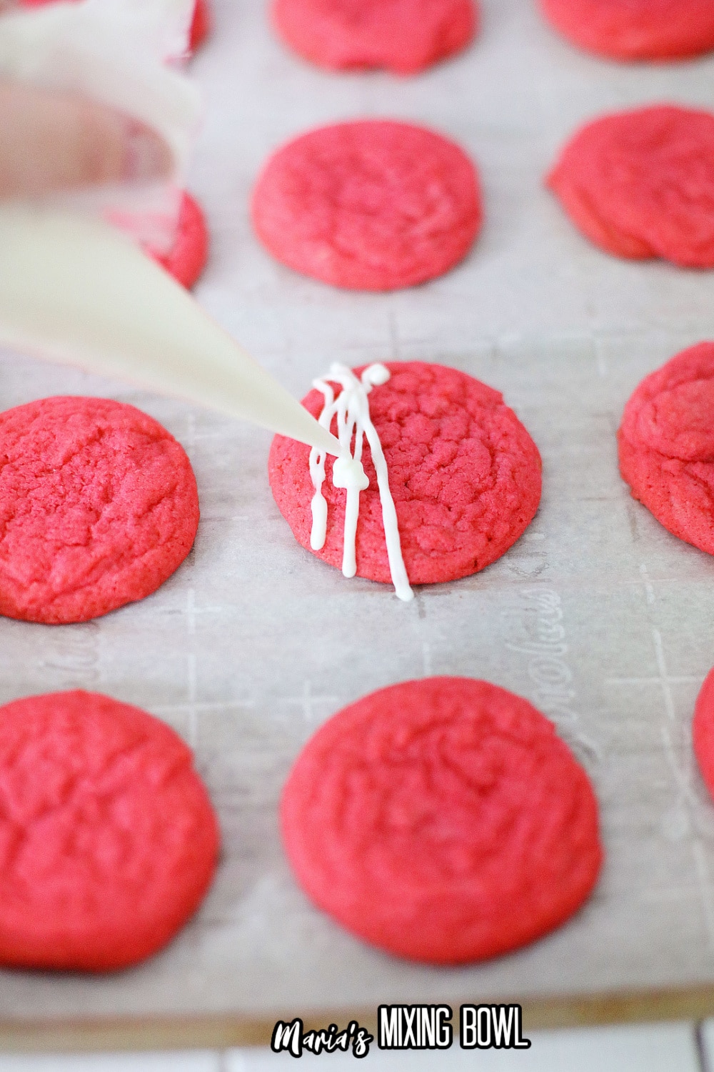 pink velvet cookies on a parchment lined baking sheet being drizzled with white chocolate
