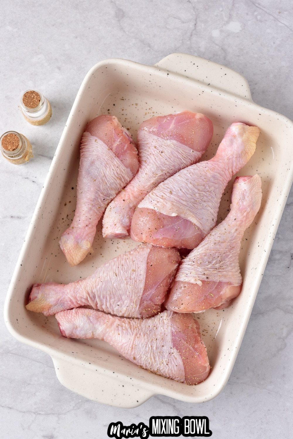 six raw chicken legs in a white ceramic baking dish being seasoned