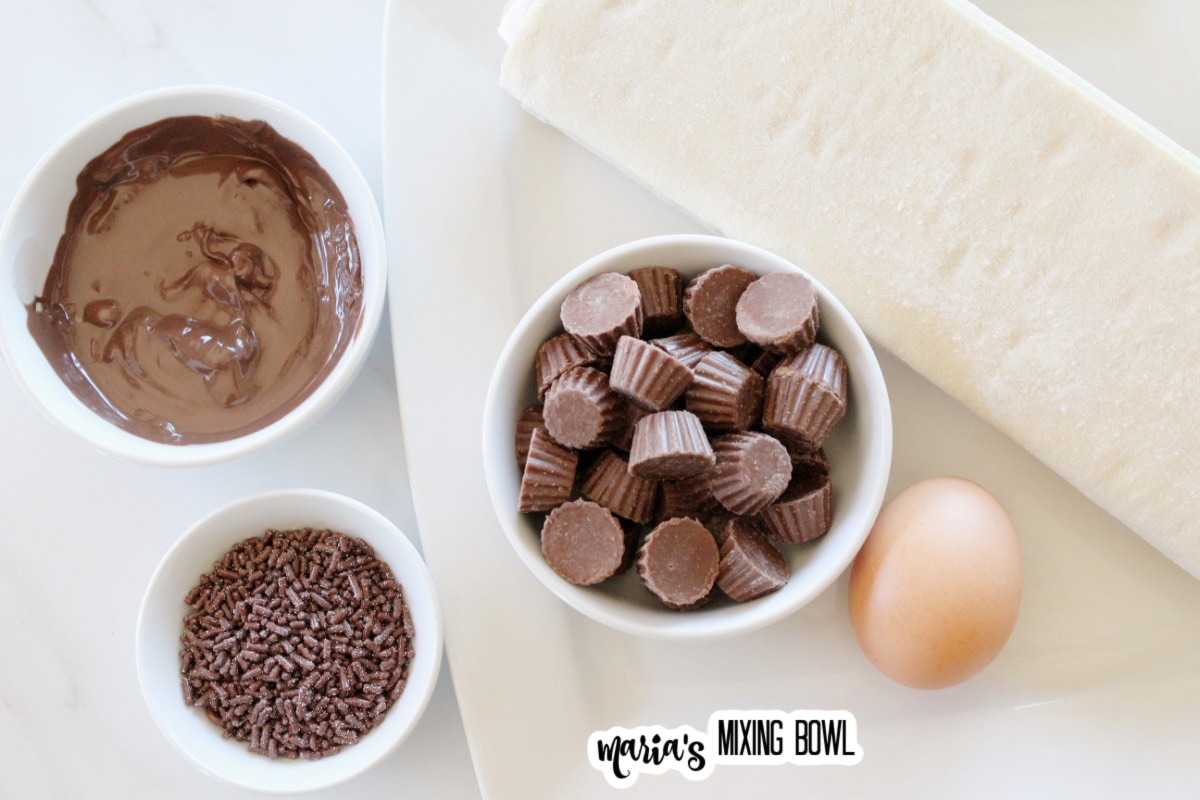 roll of puff pastry, peanut butter cups, egg, sprinkles, and melted chocolate on a white background