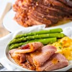close up of slices of hot honey ham on a white plate with asparagus and au gratin potatoes