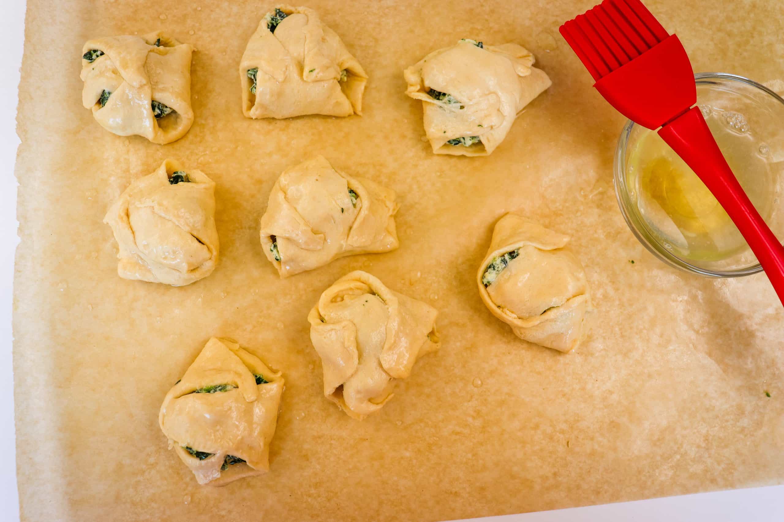 egg wash being brushed on top of spinach cheese crescent rolls on a parchment lined baking sheet