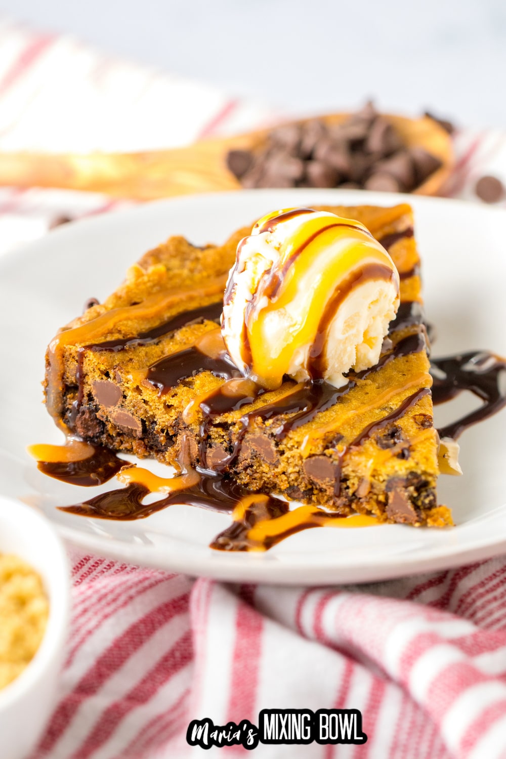 slice of cookie cake on a white plate topped with a scoop of vanilla ice cream and caramel and chocolate drizzle