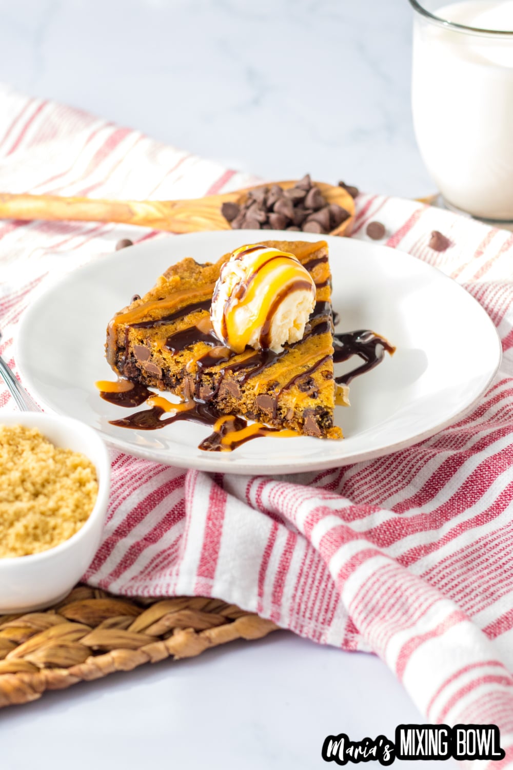 slice of cookie cake on a white plate topped with a scoop of vanilla ice cream and caramel and chocolate drizzle