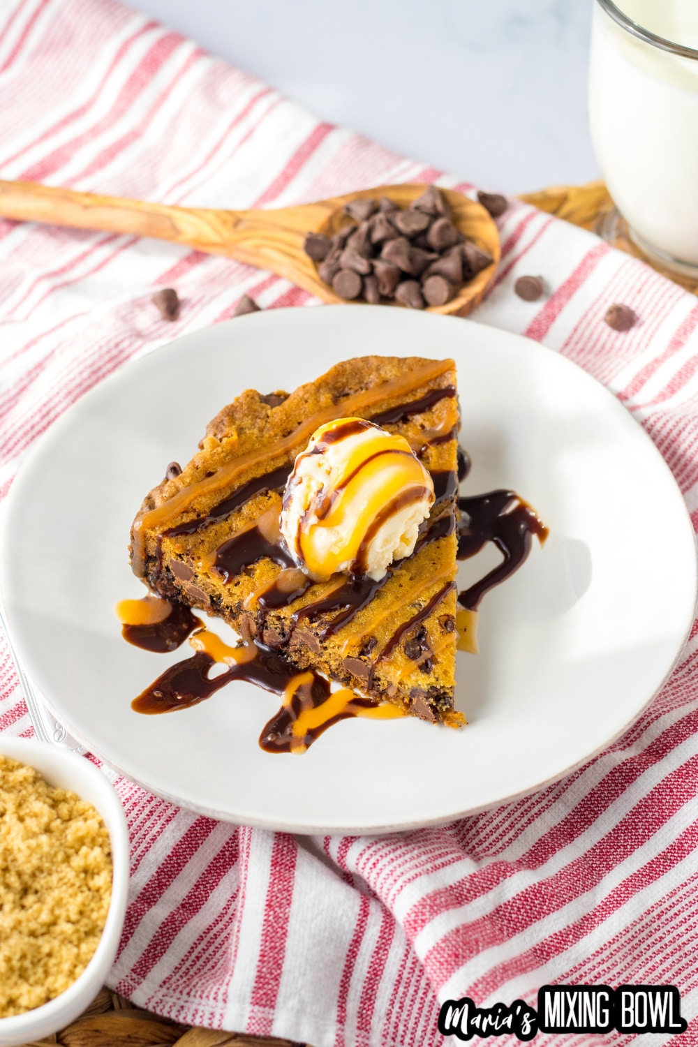 slice of cookie cake on a white plate topped with a scoop of vanilla ice cream and caramel and chocolate drizzle