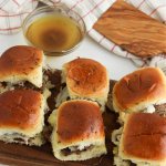 Six italian beef sliders on a wooden cutting board with a bowl of au jus in the background.