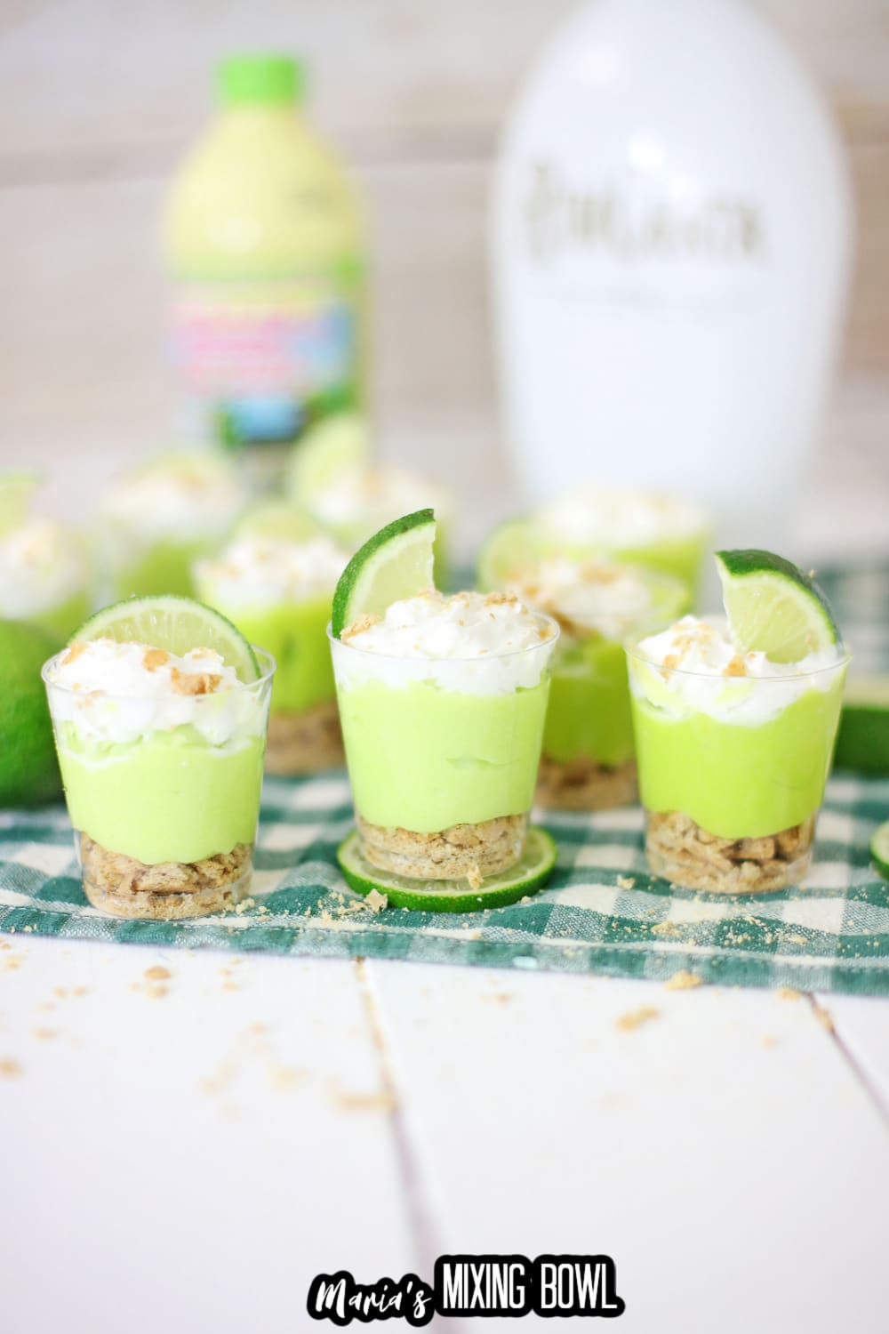 key lime pie shots on a green and white napkin with a bottle of lime juice and rum chata in a background