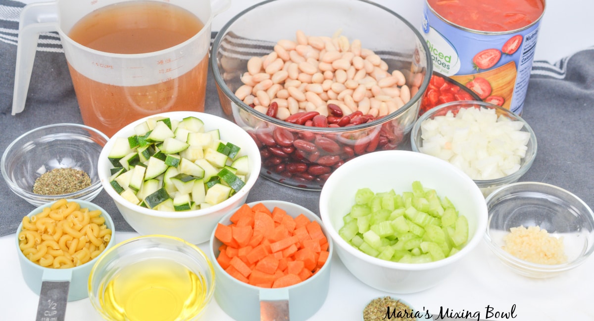 broth, tomatoes, beans, seasonings, onion, celery, zucchini, carrots, garlic, oil, and pasta laid out on a counter