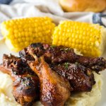 four chicken drumsticks on top of a bed of mashed potatoes with corn cobs in the background