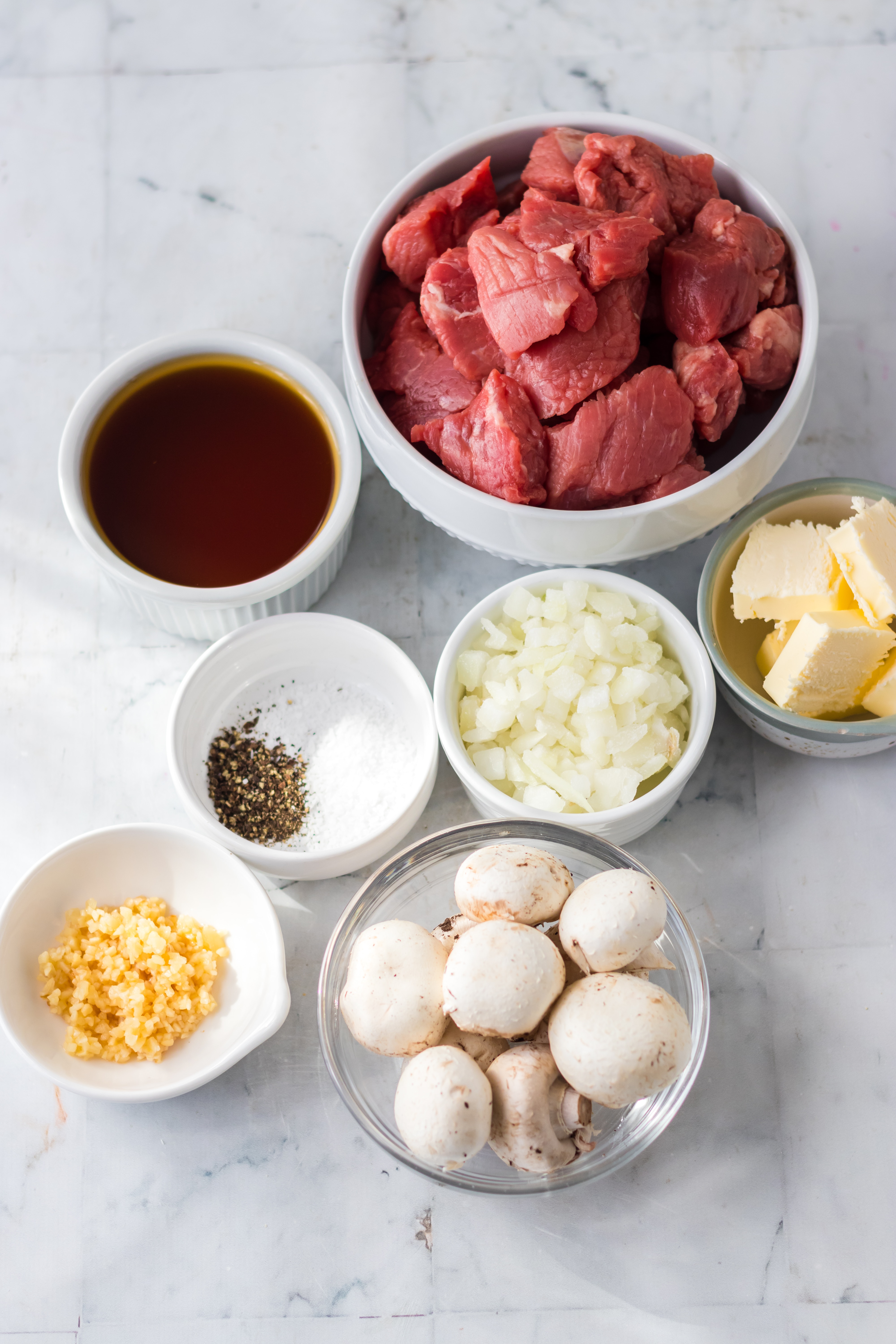 steak bites, garlic, onions, seasoning, butter, and mushrooms on a white marble background