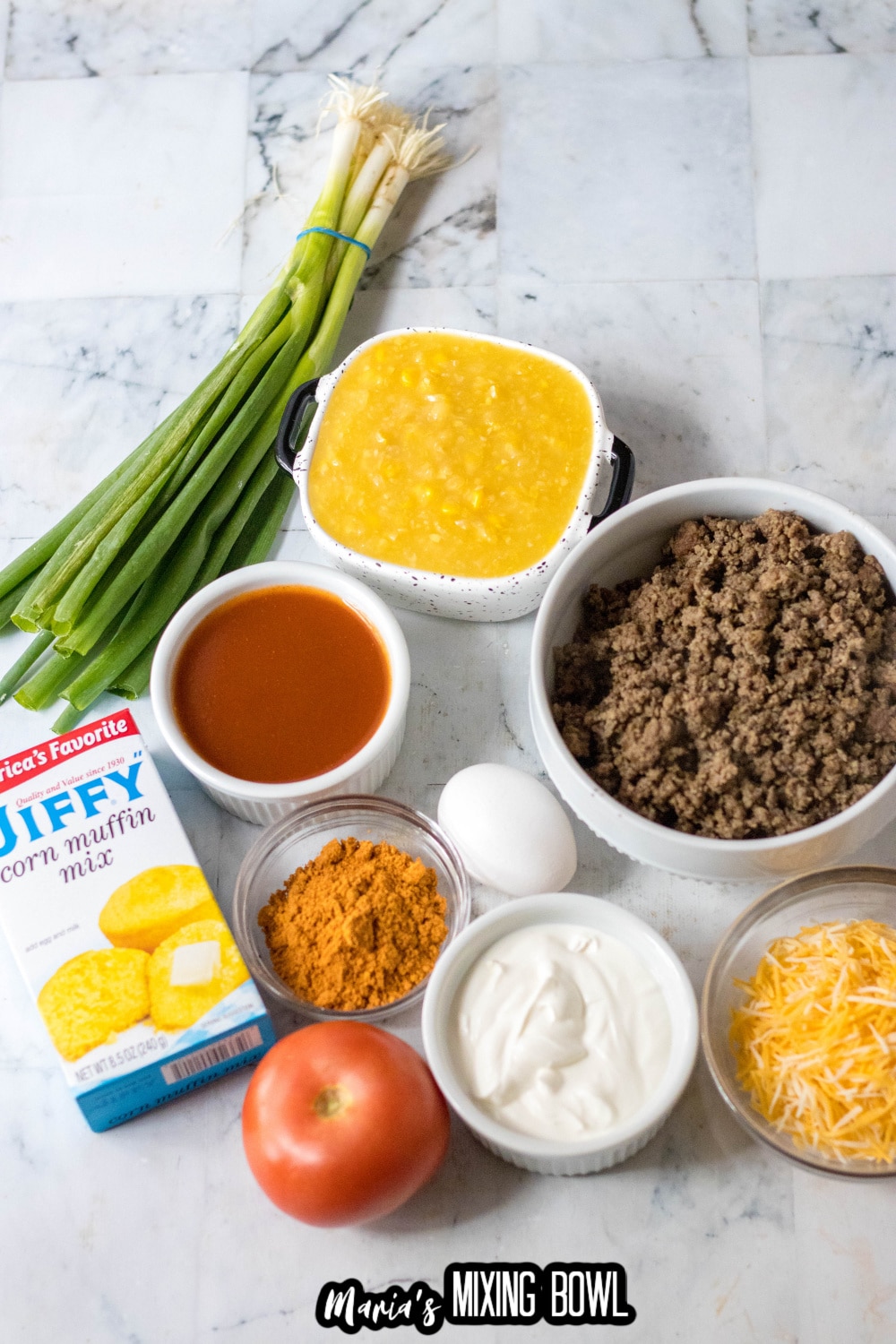 green onions, creamed corn, ground beef, jiffy mix, taco seasoning, tomato, egg, sour cream and cheese on a marble backdrop