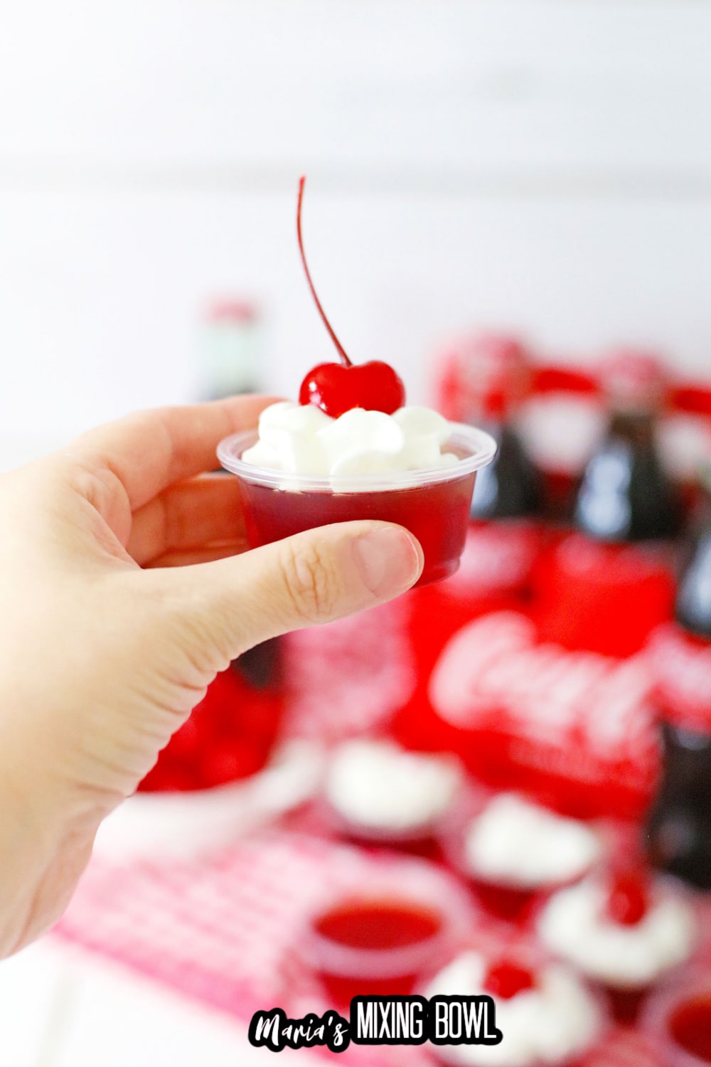 cherry jello shot topped with whipping cream and a cherry held in a woman's hand