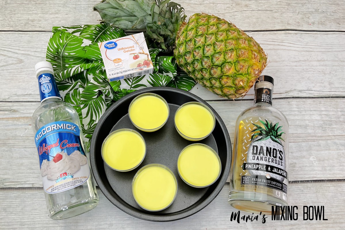 five shots of dole whip in a cake pan, surrounded by bottles of alcohol, gelatin, and pineapple