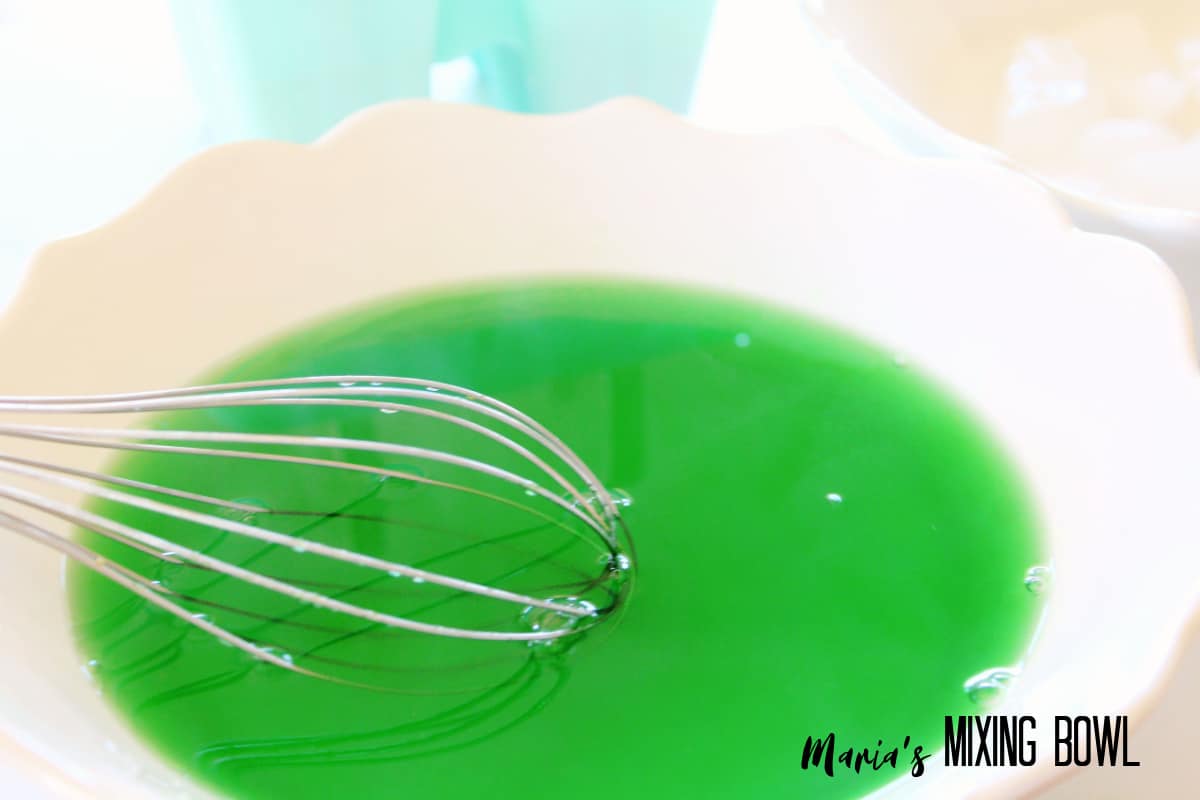 green gelatin being mixed with a whisk in a white bowl