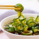 cucumber salad in a white bowl with a stack of cucumbers behind it