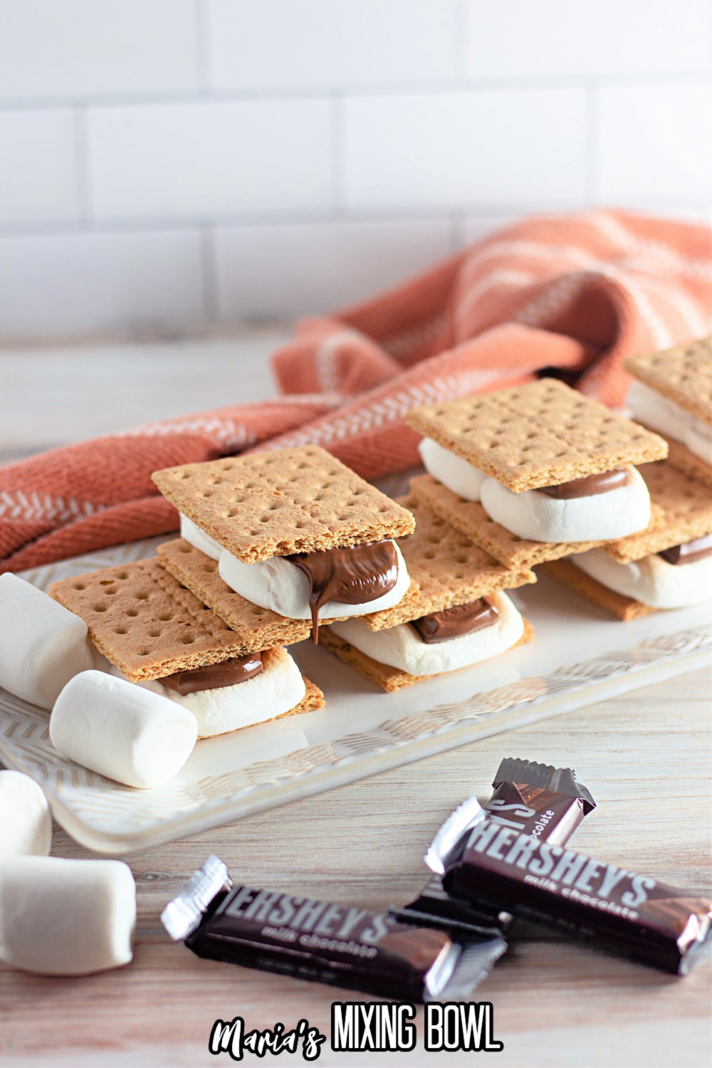 six smores on a white platter surrounded by chocolate bars and marshmallows