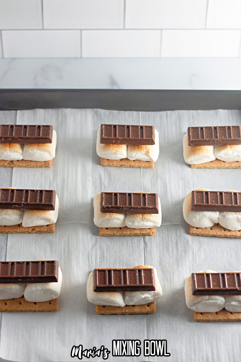 chocolate being placed onto toasted marshmallows and graham crackers on a parchment lined baking sheet