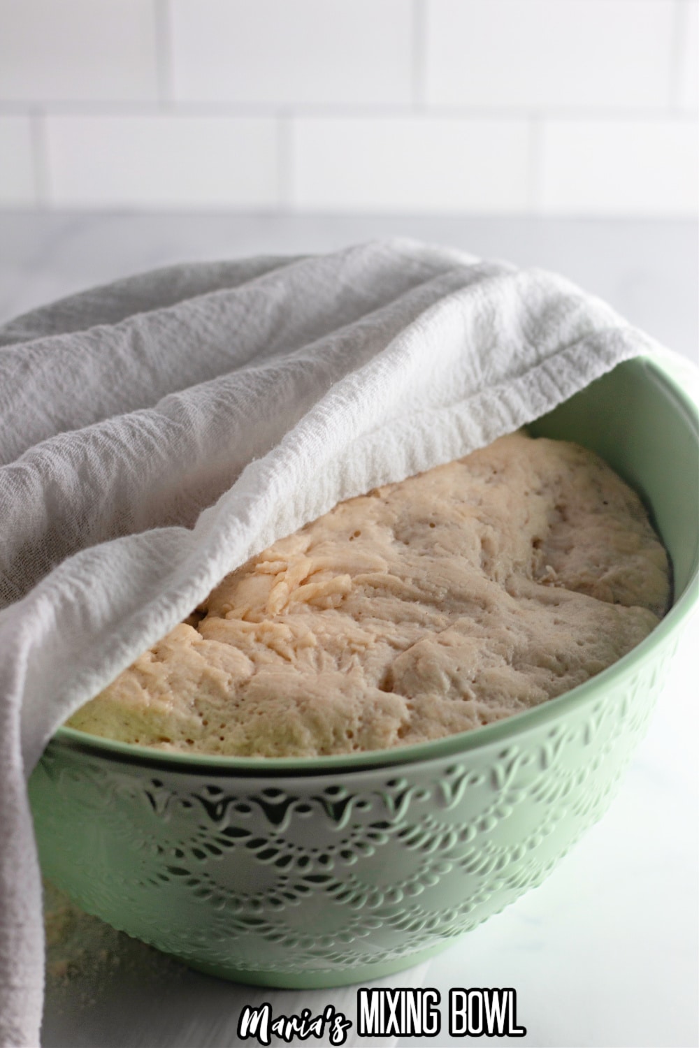 a green bowl filled with proofed bread dough and covered with a flour sack towel