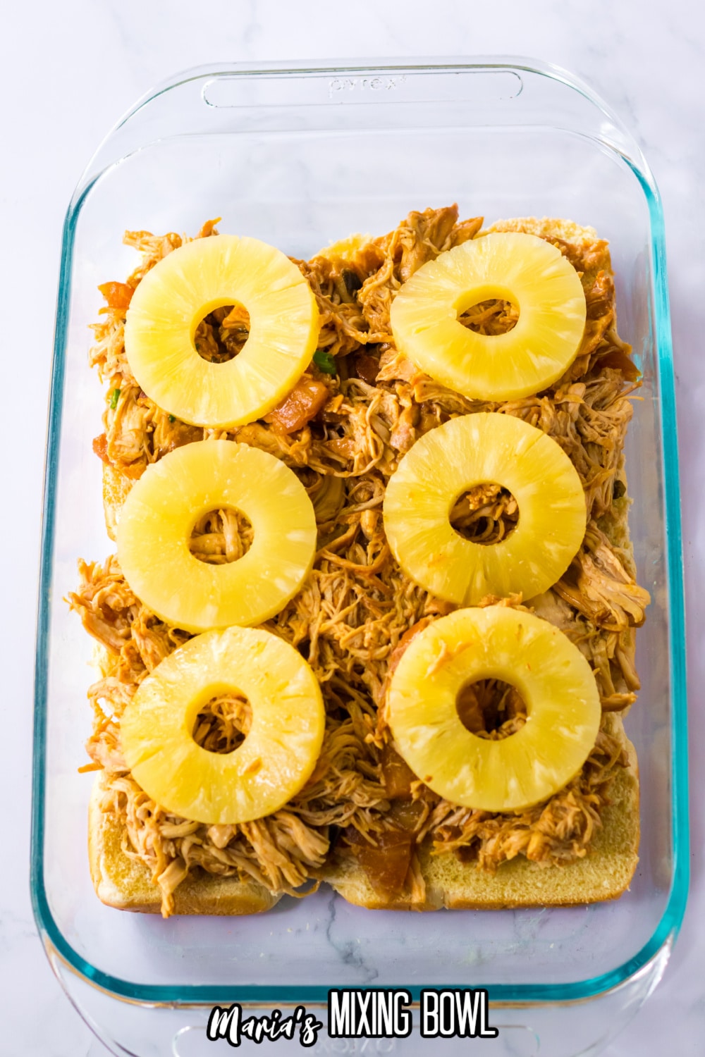 hawaiian rolls topped with shredded chicken and pineapple slices in a glass baking dish