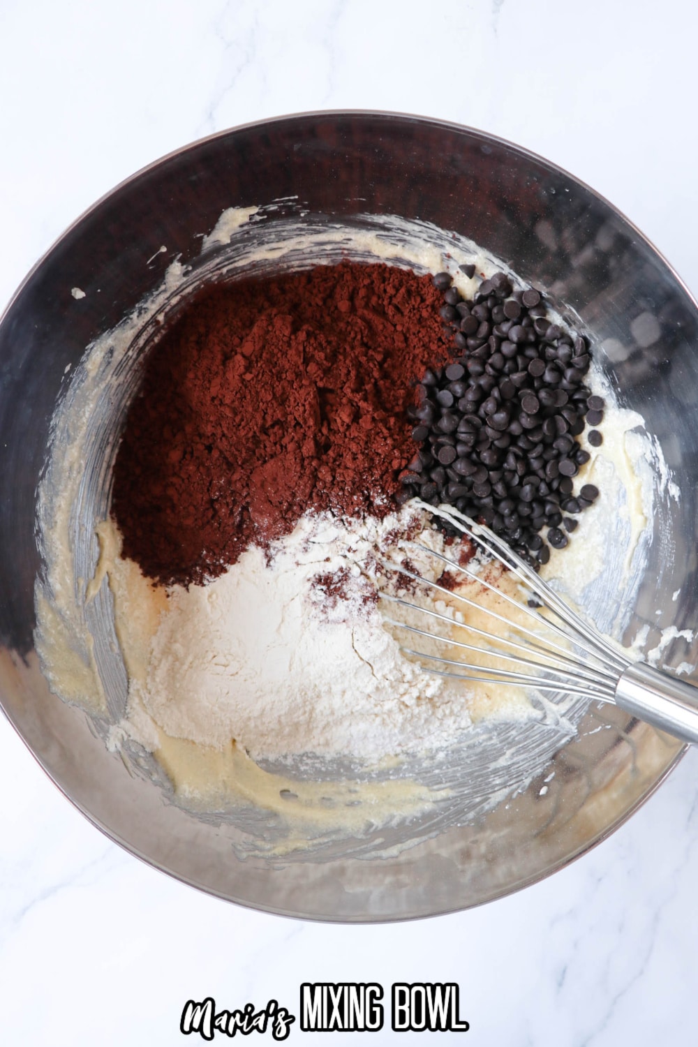 flour, sugar, cocoa powder, and chocolate chips being mixed in a stainless steel mixing bowl