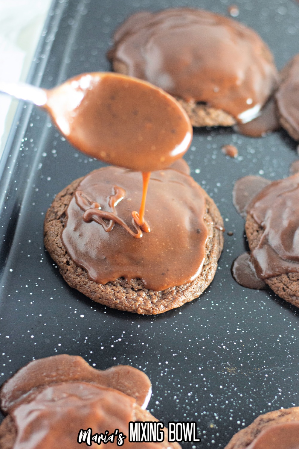 chocolate glaze being poured on top of cookies on a dark baking sheet