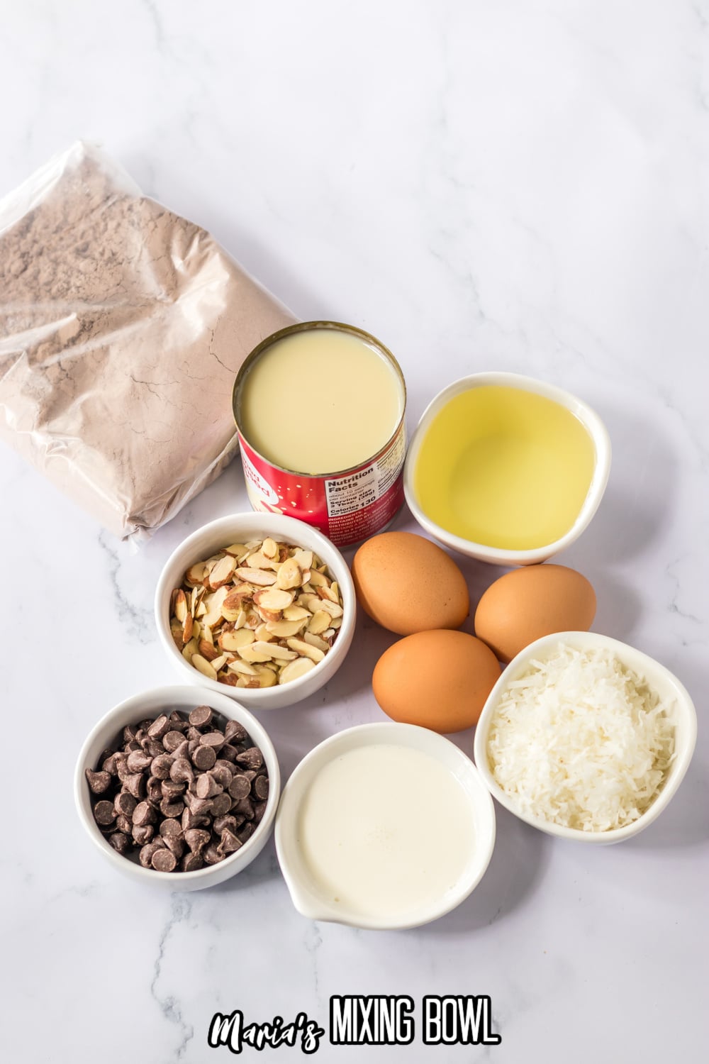 cake mix, can of condensed milk, oil, sliced almonds, chocolate chips, heavy cream, eggs, and shredded coconut on a white background