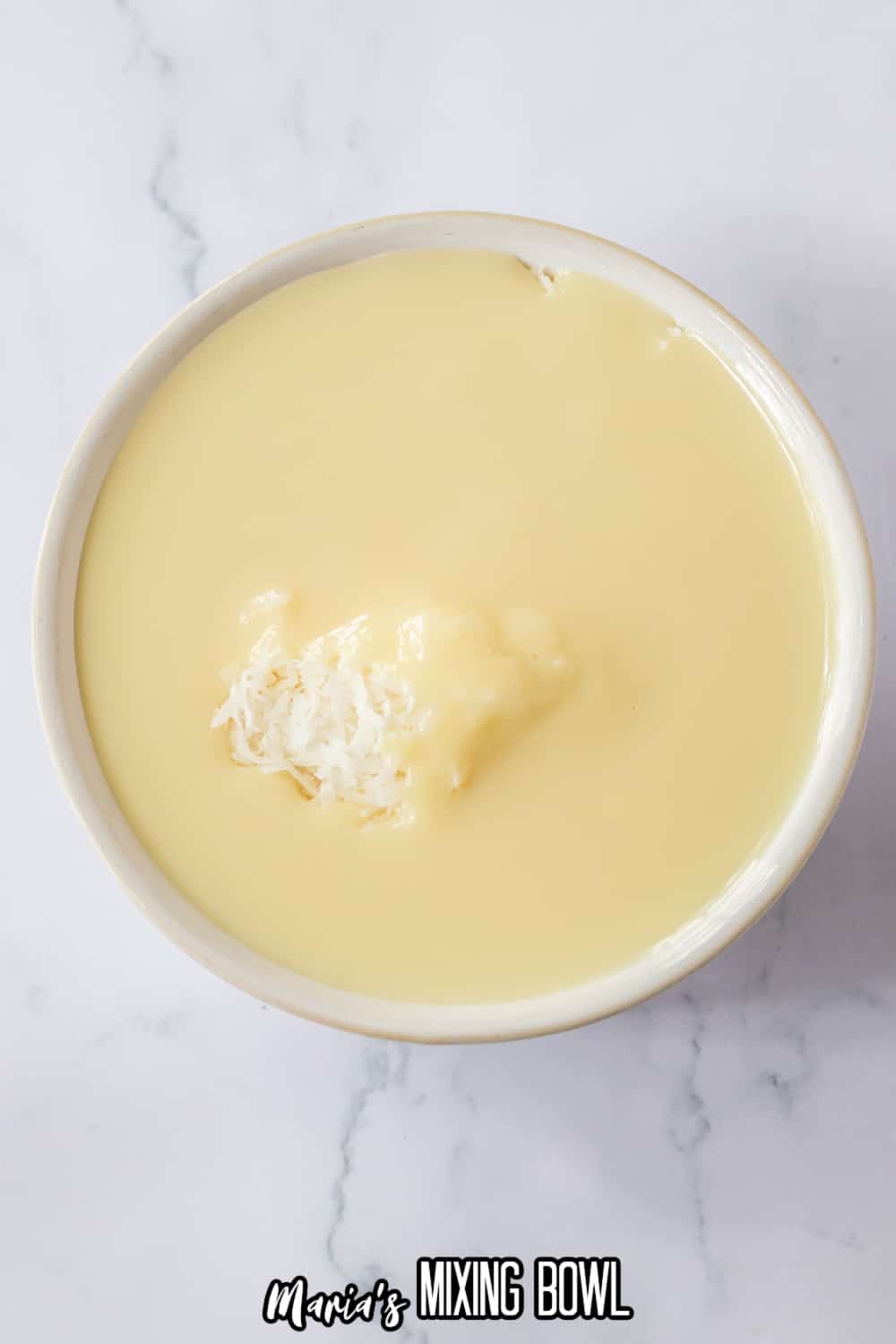 shredded coconut being mixed into a white bowl filled with sweetened condensed milk