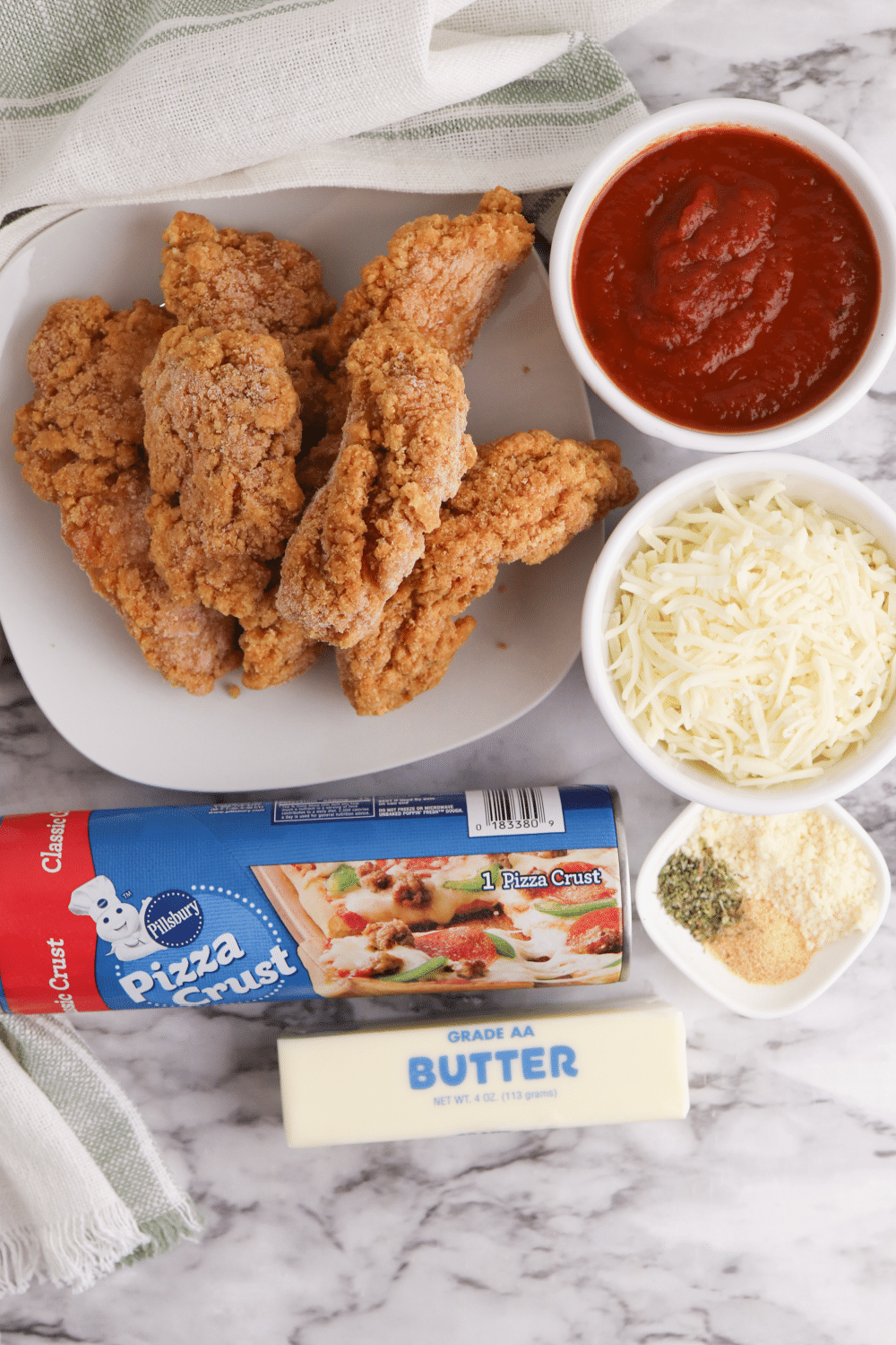 plate of chicken tenders, small bowl of pizza sauce, small bowl of mozzarella cheese, can of pizza crust, stick of butter, bowl of seasonings and parmesan cheese