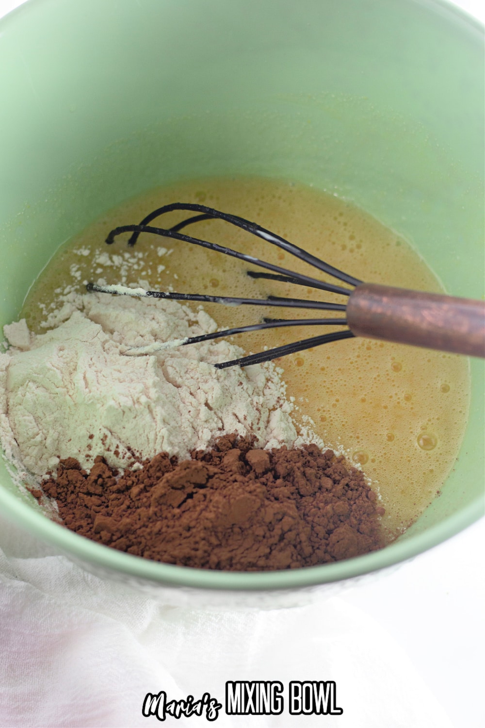 flour and cocoa powder being added to the wet ingredients in a green mixing bowl